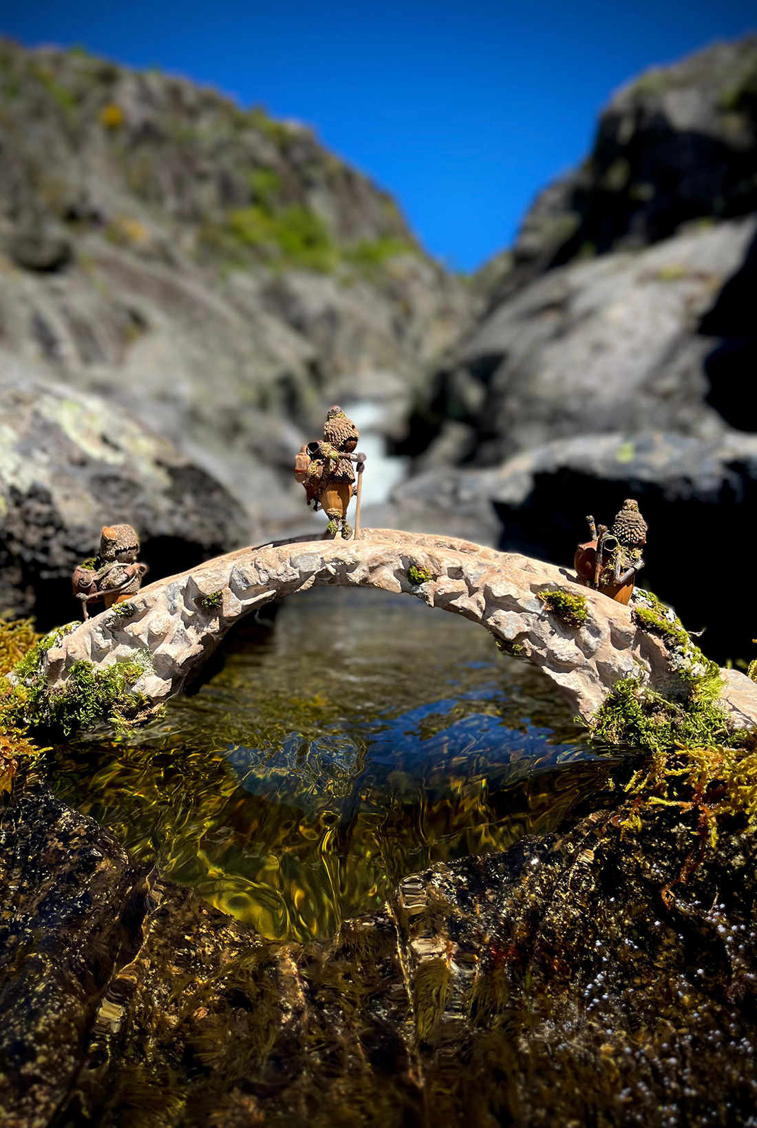 Tres figuras, Los Bellotines, elaboradas con bellotas y otros elementos naturales en el bosque como si fueran senderistas pasando un puente.