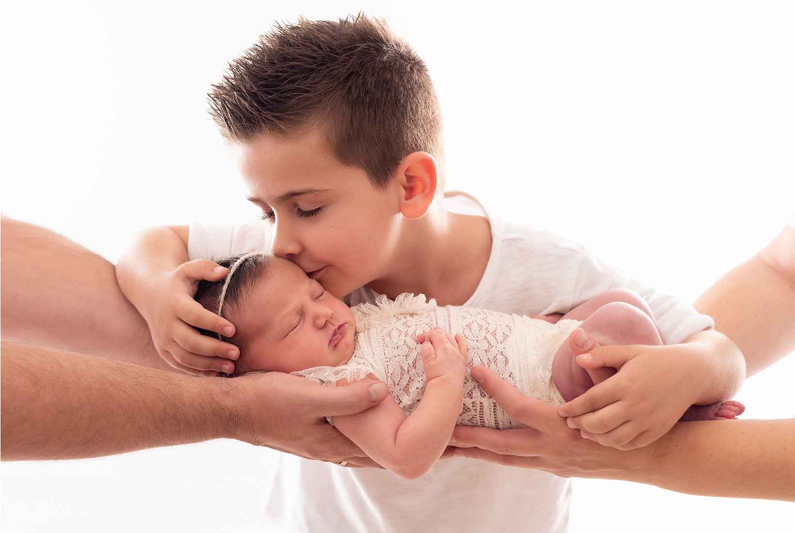 Fotografía de un bebés sobre los brazos de sus padres mientras su hermano le da un beso. De Jennifer Suárez Fotografía.